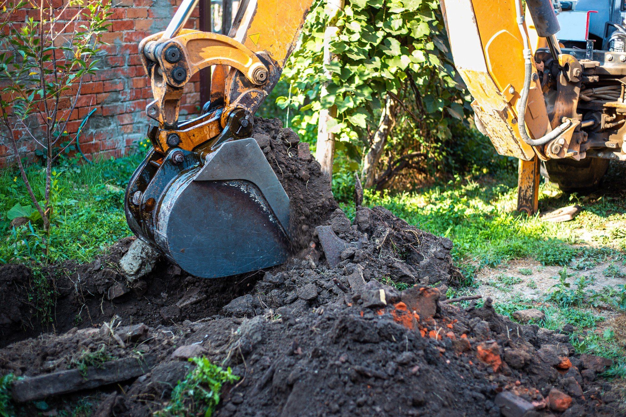 Backhoe bucket digging a trench in a garden beside a brick wall